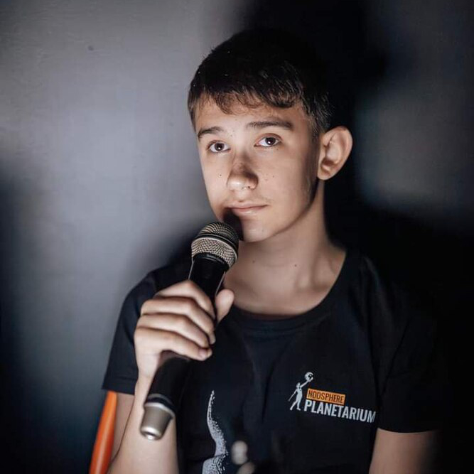 Maksym, a young autistic man from Ukraine, is holding a microphone. He wears a t-shirt that says "Noosphere Planetarium".